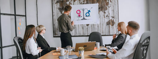 Hombre señala un cartel con símbolos de género frente a un grupo de personas sentadas en una mesa de reunión